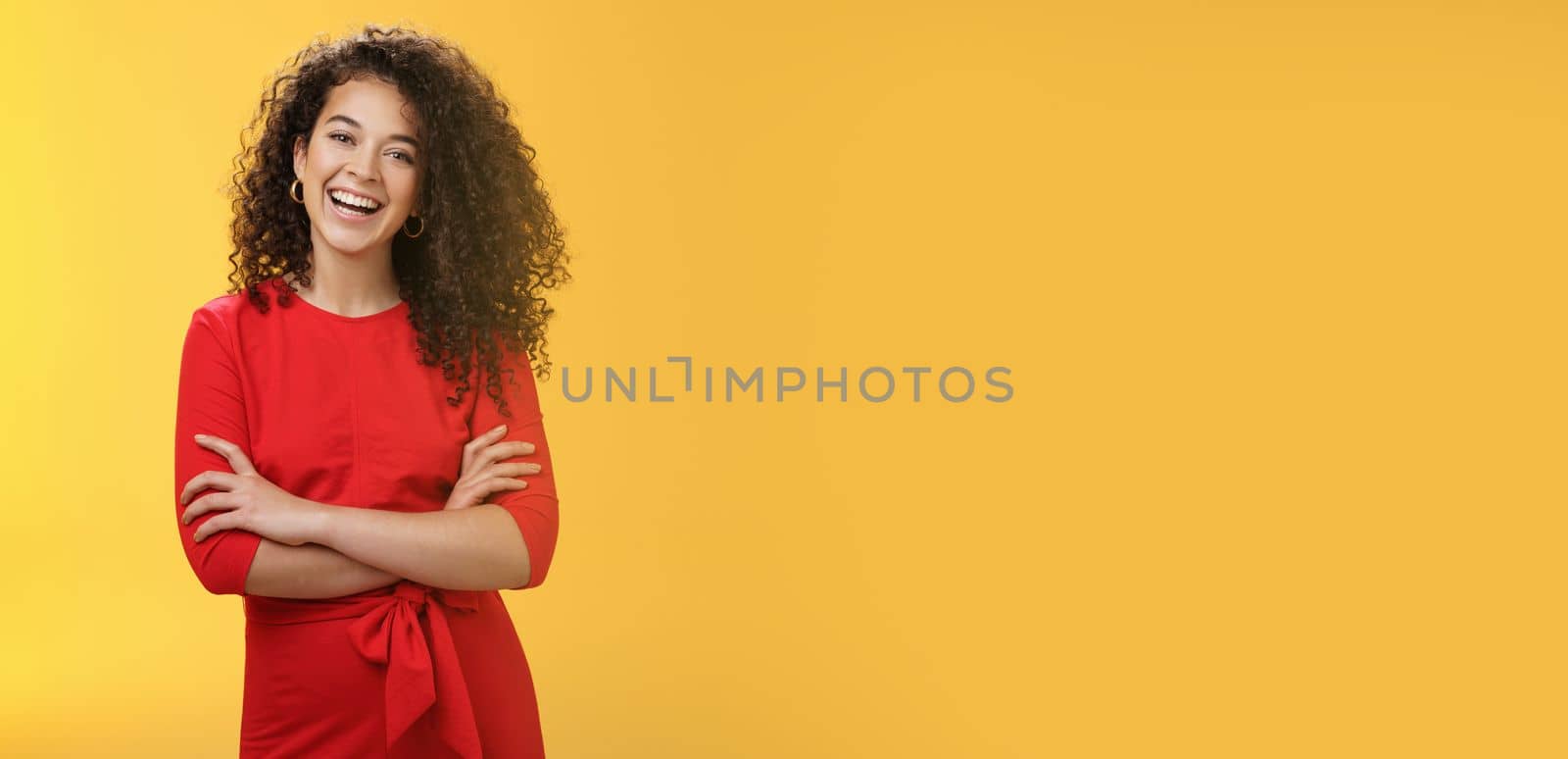 Self-assured happy enthusiastic curly-haired female reporter in cute red dress laughing carefree, having fun tilting head amused and holding hands crossed over body in confident pose over yellow wall by Benzoix