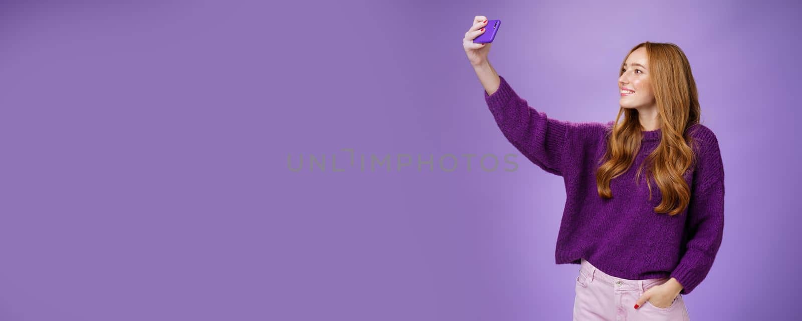 Attractive confident and stylish happy 20s woman with ginger hair standing carefree with joyful smile over purple background extending hand with smartphone as taking selfie happily. Technology and lifestyle concept