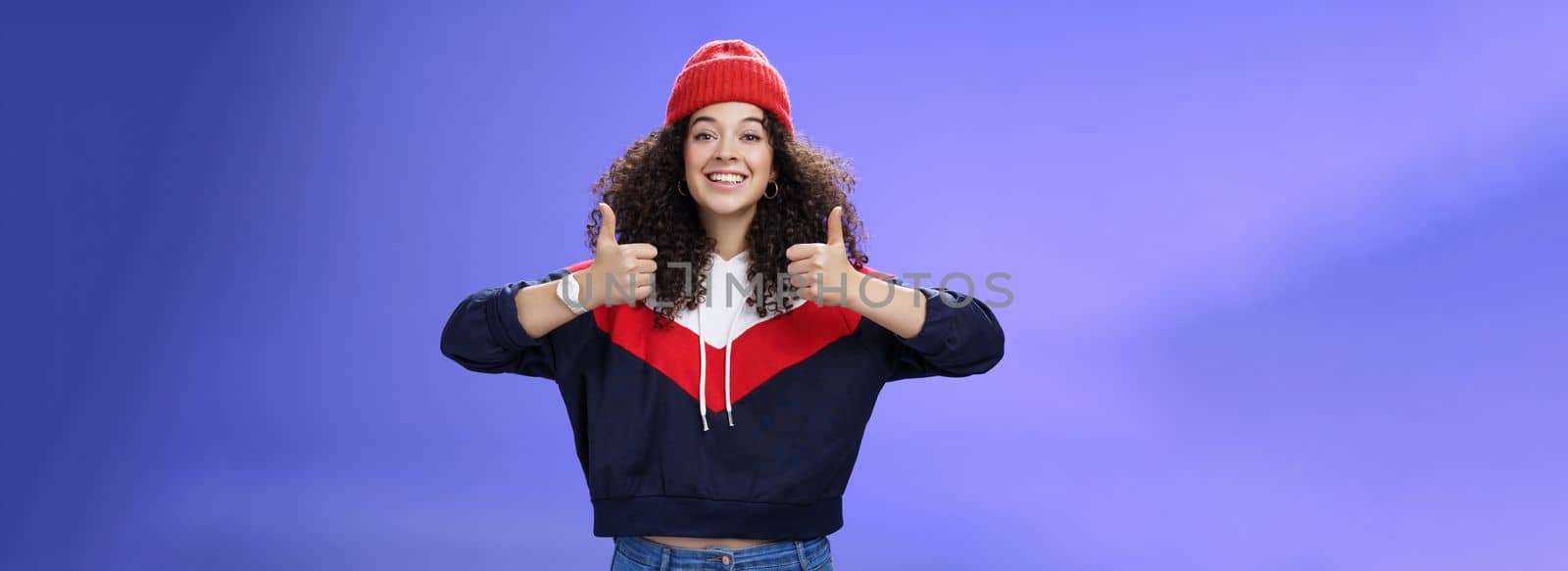 Girl totally agrees recommends cool place showing thumbs-up and smiling excited with happy grin standing upbeat and delighted against blue background in warm beanie and sweatshirt by Benzoix
