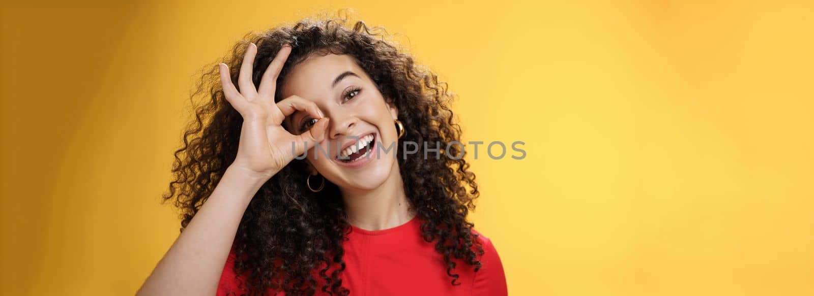 Hey I see you. Charismatic funny and playful cute tender girlfriend with curly hair showing okay or zero gesture over eye peeking at camera and smiling broadly, tilting head right over yellow wall.
