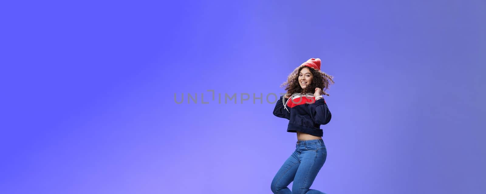 Studio shot of cute girl with curly hair in beanie sweatshirt and sneakers jumping playful and carefree over blue background, having fun enjoying cool weather smiling broadly as flying in air.