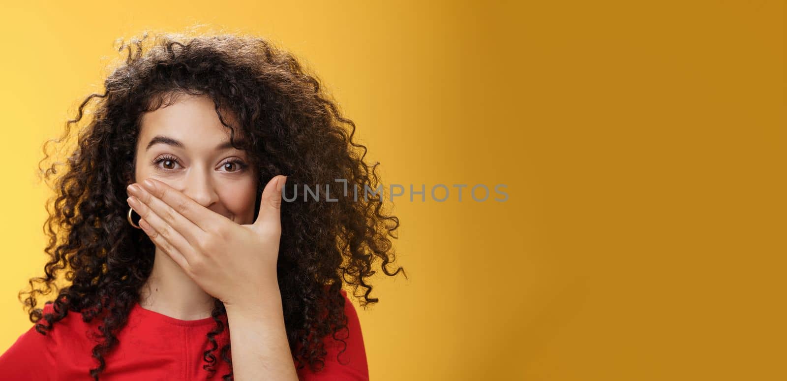 Close-up shot of excited and happy attractive female with curly hair giggling, chuckling and covering mouth as smiling standing amused and joyful as mocking friend over yellow background by Benzoix