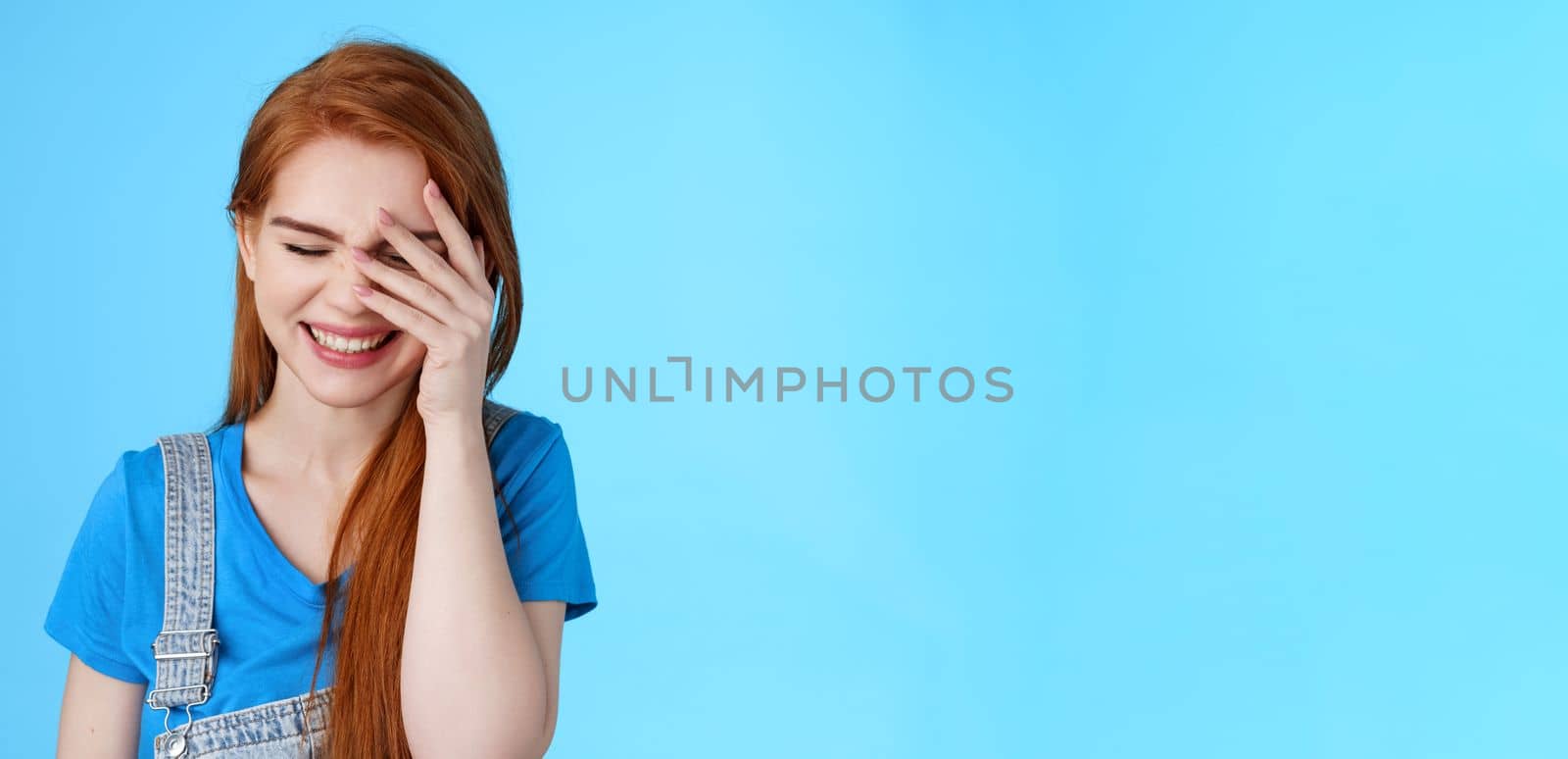 Close-up carefree cheerful good-looking redhead woman laughing, blushing cute sincere giggle, bend head down close eyes smiling broadly, chuckling funny joke, stand blue background. Copy space