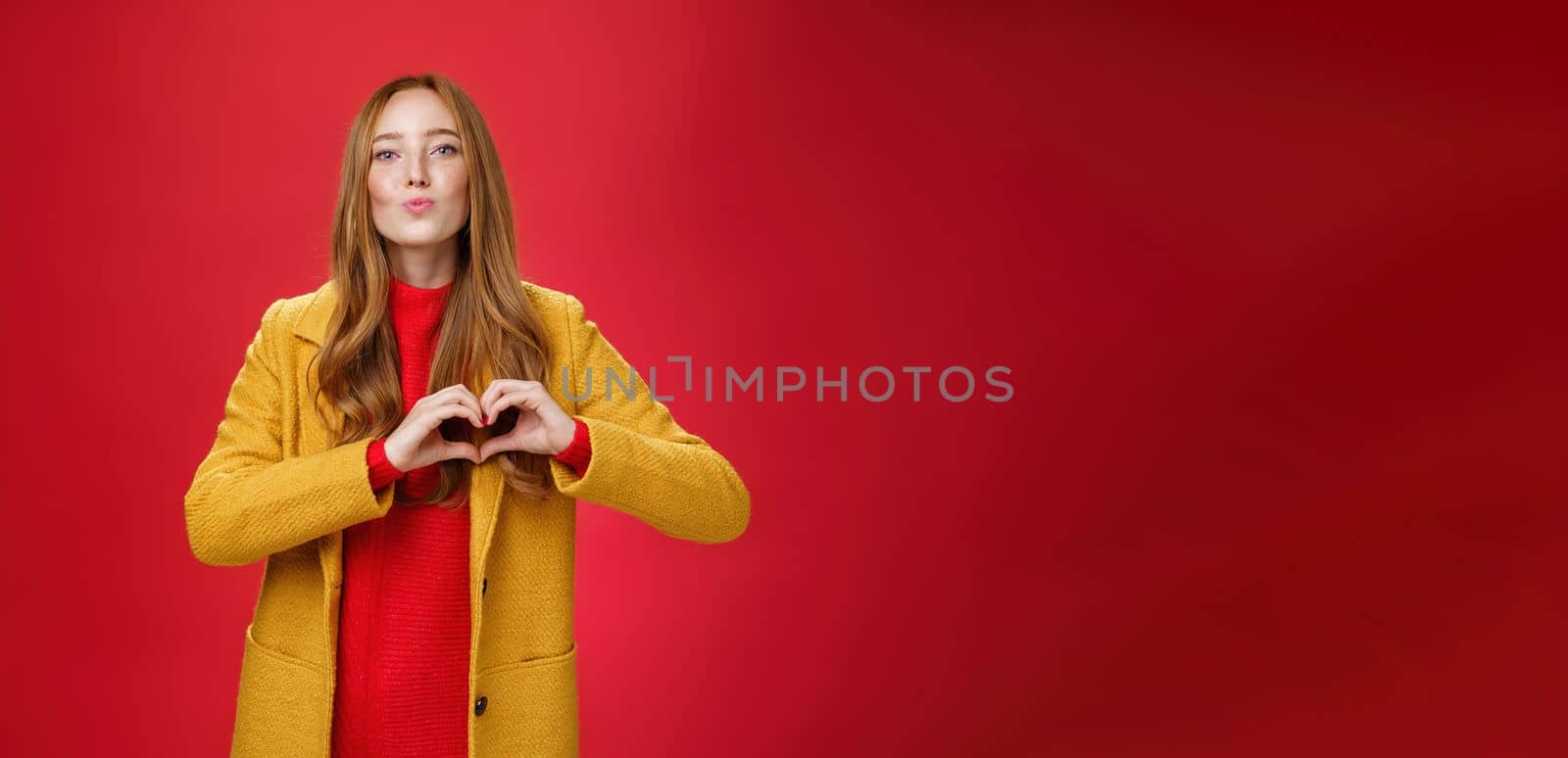 Love you all. Portrait of romantic and stylish good-looking flirty redhead female with freckles and blue eyes folding lips to give kiss showing heart gesture, confessing in sympathy over red wall.