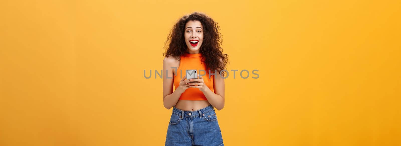 Indoor shot of surprised happy and delighted attractive european female with curly haircut in cropped top holding smartphone gazing at camera amazed and pleased receiving awesome news via messages. Technology concept