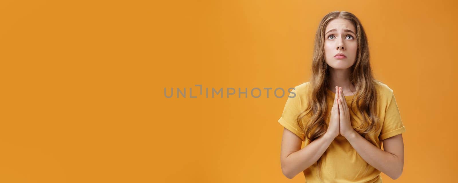 Hopeful uneasy and worried gloomy faithful girl in t-shirt holding hands in pray against chest looking up with sad look praying making wish for good well of family posing over orange wall. Body language concept