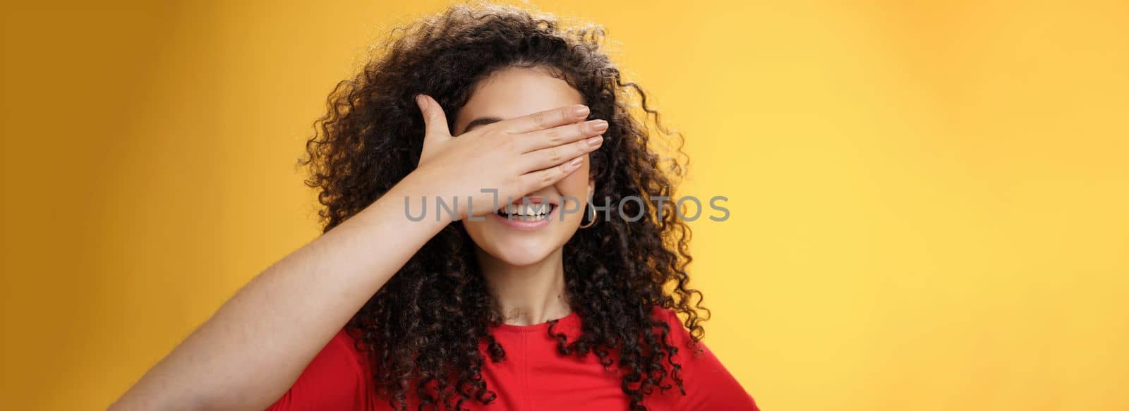 Close-up shot of dreamy happy young cute woman with curly hair covering eyes with palm as counting or playing peekaboo smiling broadly anticipating surprise happen over yellow background by Benzoix