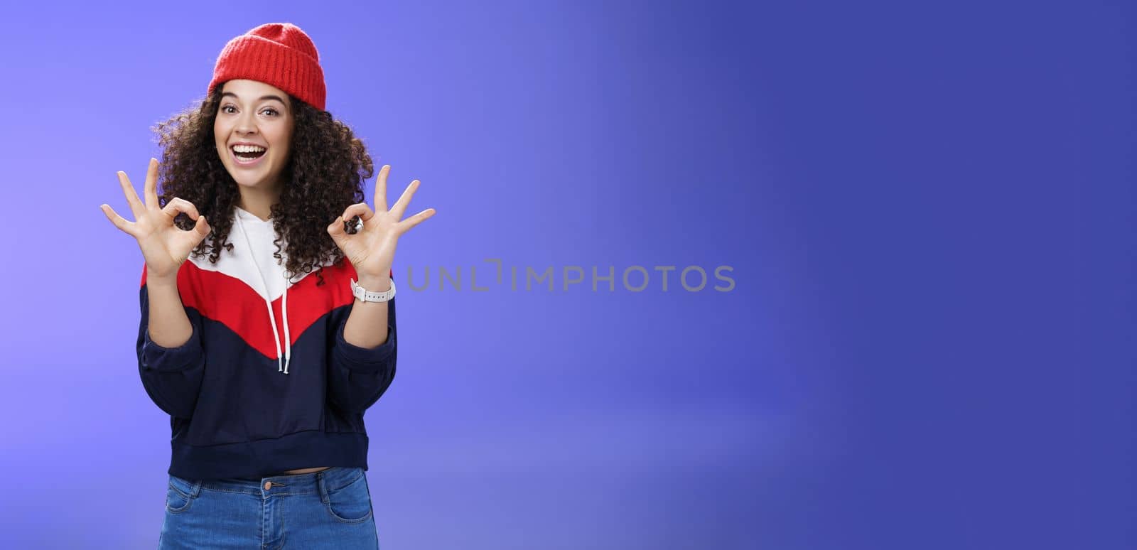 Got under control. Portrait of happy charming smiling curly-haired female in warm winter hat and sweatshirt smiling broadly and showing okay or excellent gesture as approving, liking cool movements.