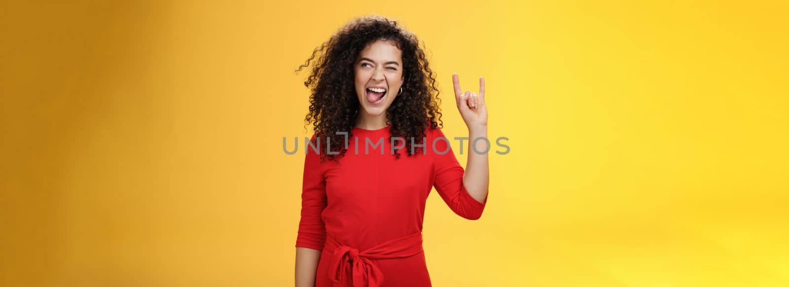 Rebellious meloman leaving in tender girl. Thrilled and carefree curly-haired woman in red dress sticking out tongue and looking right with smile as showing rock-n-roll gesture, enjoying cool music.