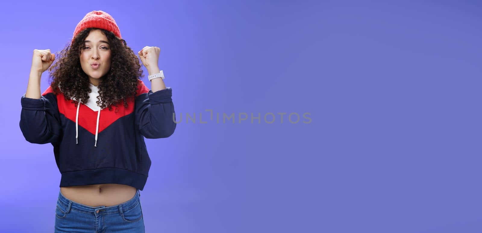 Charming young 20s girl with curly hair in warm beanie raising hands as belly showing under sweatshirt triumphing, celebrating awesome win folding lips from joy and excitement over blue background.