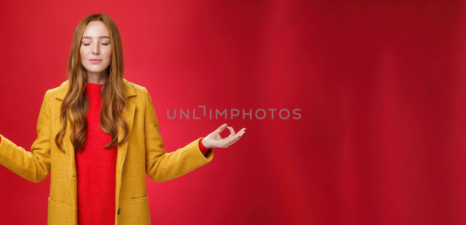 Girl keep calm releasing stress with meditation, posing in yellow coat, close eyes and looking relieved as extending hands sideways with mudra gesture, doing yoga against red background in lotus pose.