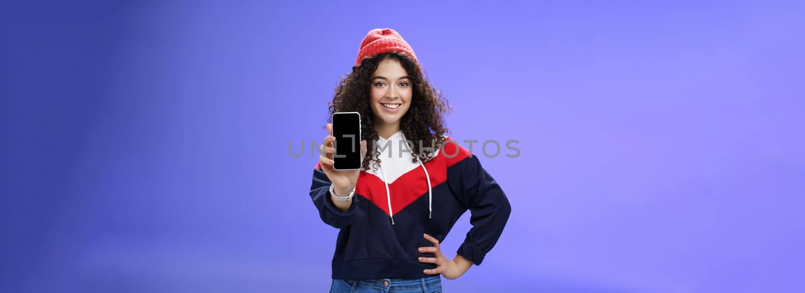 Girl showing her post as pulling smartphone screen towards camera smiling delighted waiting opinion, holding hand on waist in confident pose wearing warm winter red beanie and sweatshirt by Benzoix