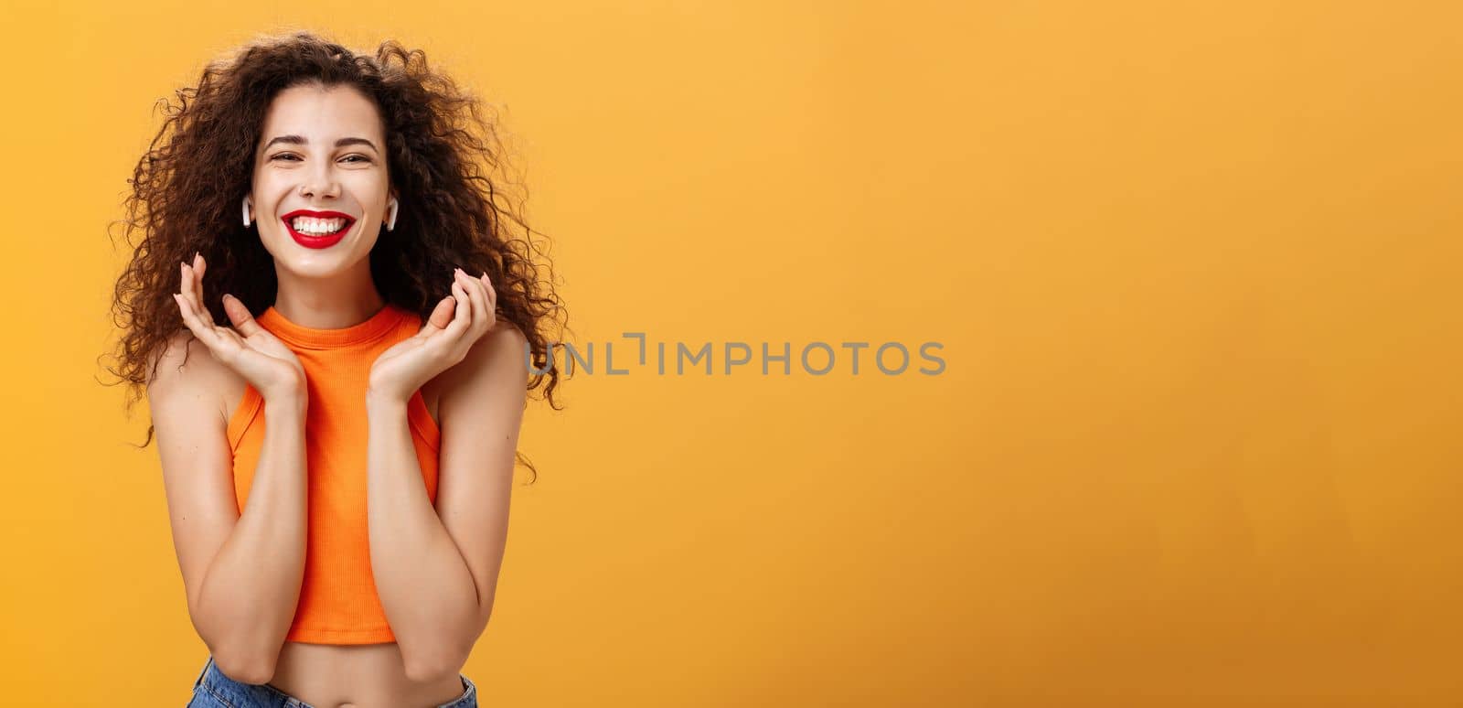 Stylish happy attractive urban female with curly hairstyle. and nose ring smiling broadly feeling delighted listening favorite track in wireless earbuds raising palms near face posing over orange wall.