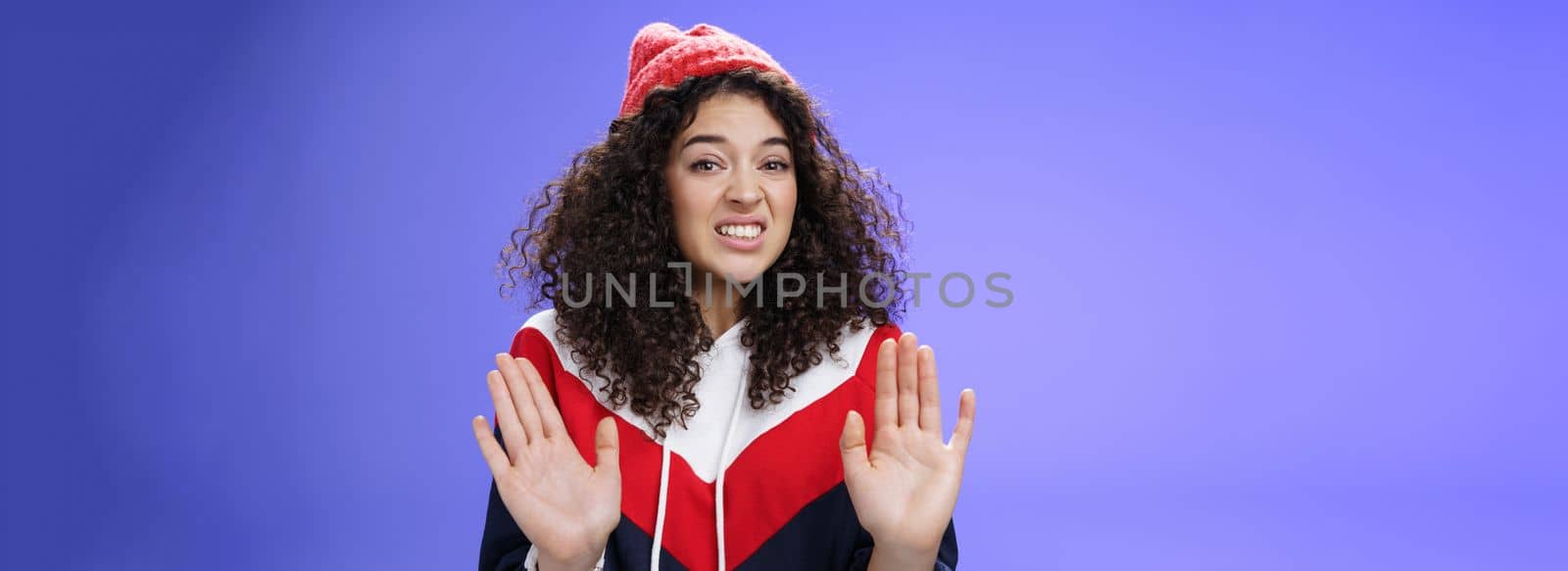 Displeased european woman with curly hair raising palms near chest and waving in refusal and rejection gesture grimacing unsatisfied and unwilling to participate in suspicious action over blue wall by Benzoix