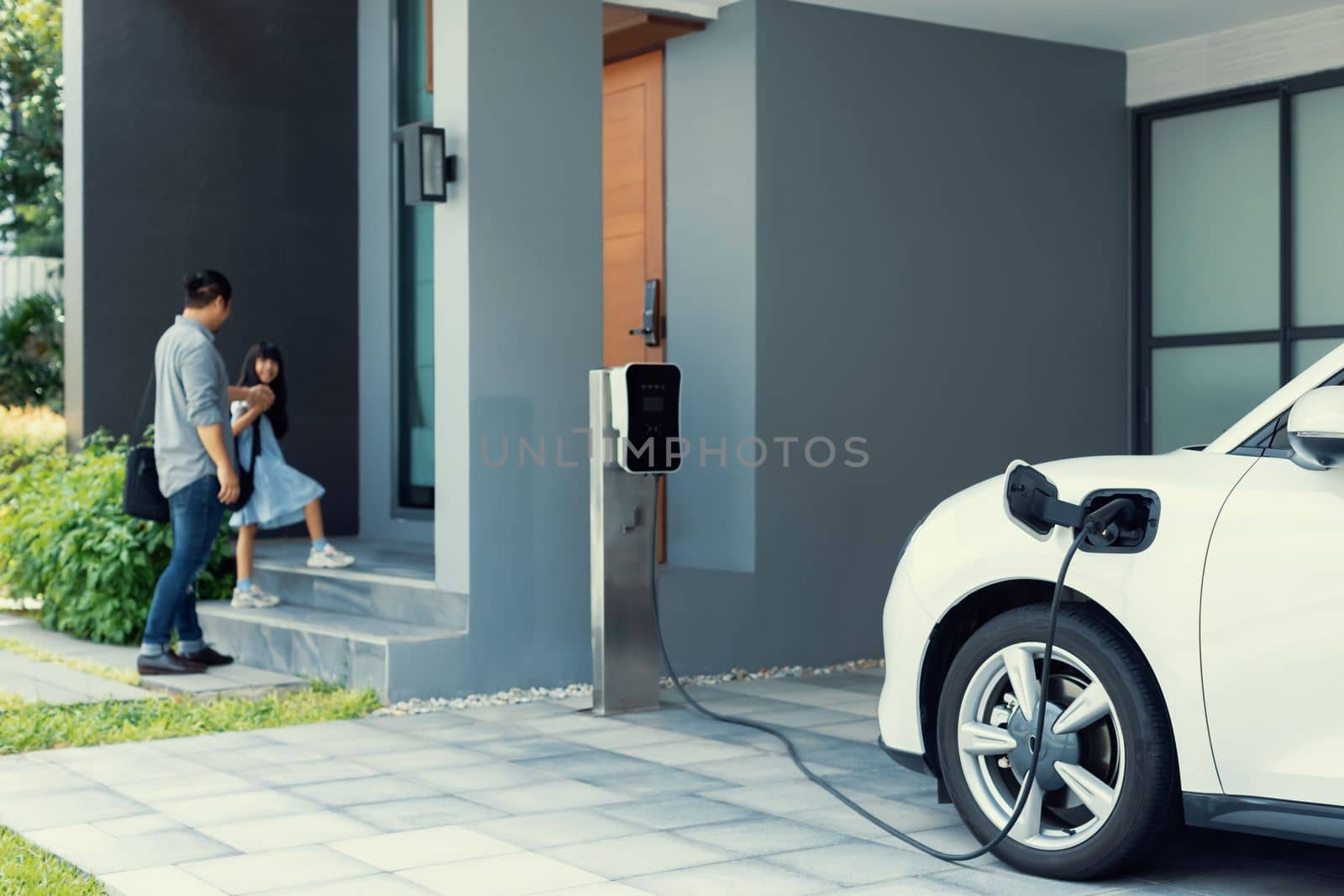Focus progressive electric vehicle recharging at home charging station using clean and renewable energy with blurred father and daughter walking in background for future renewable energy concept.