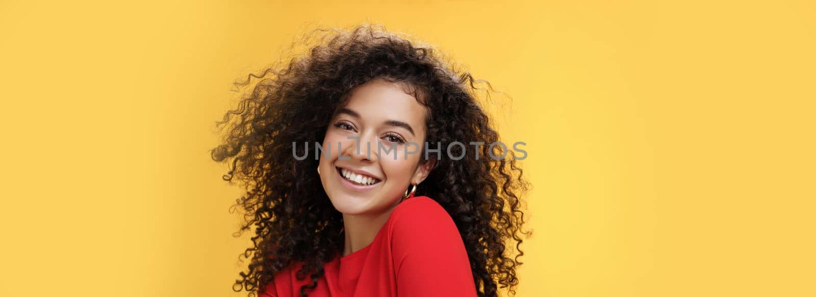 Romantic and flirty young cute caucasian girlfriend with curly hair in red blouse making silly pose smiling joyfully and lifting shoulder coquettish smiling broadly at camera over yellow wall by Benzoix
