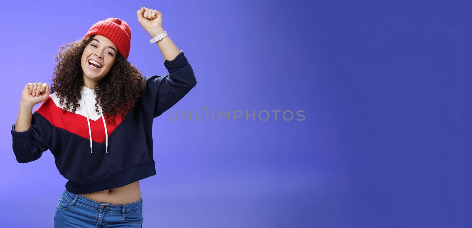 Hey come on dance with me. Friendly-looking bright and stylish cheerful woman with curly hairstyle wearing warm beanie raising hands up as enjoying great day, having fun outdoors over blue wall.