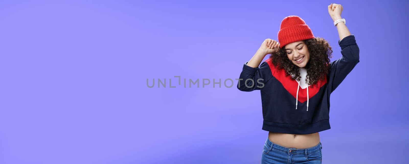 Girl celebrating favorite season in year. Portrait of carefree and joyful dancing woman with curly hair in cute red hat lifting hands in dance movements smiling enjoying music and holidays by Benzoix