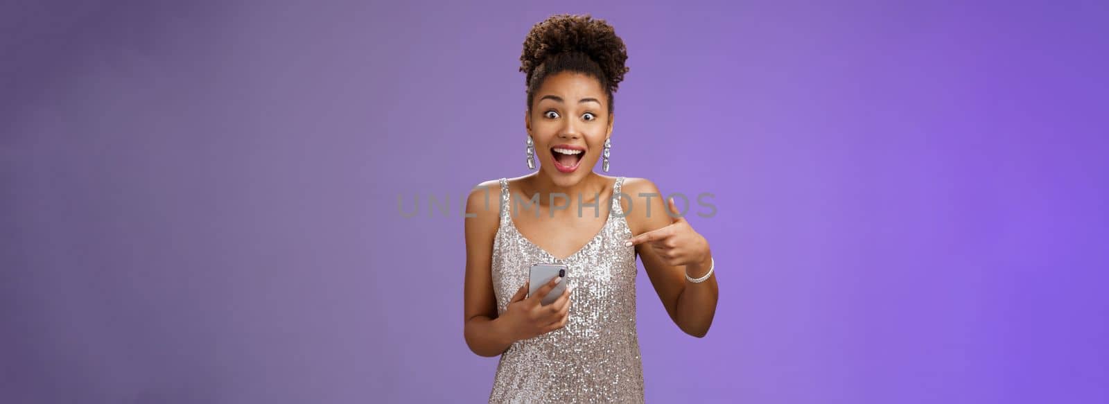 Amused charming glamour african-american woman in glittering silver dress astonished thrilled pointing smartphone receive incredible good perfect news telling you, standing blue background by Benzoix