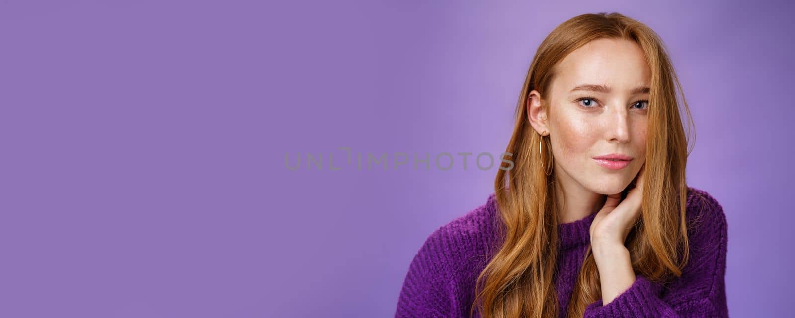 Sensual and romantic woman with red hair and freckles touching cheek gently as curls dropping on half face gazing charmed and flirty at camera, smiling being tender and coquettish over purple wall.