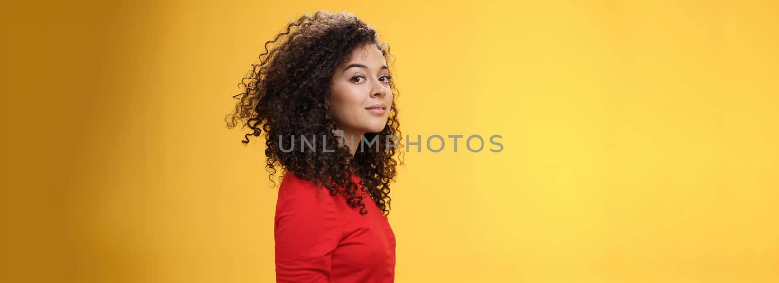 Profile shot of stylish and cool attractive curly-haired european 25s woman in red dress turning at camera and smiling broadly with delighted carefree expression, having fun over yellow background by Benzoix