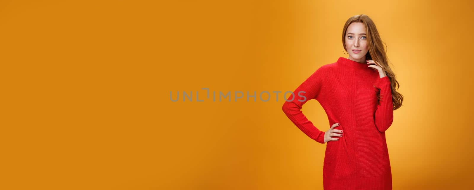 Lifestyle. Attractive sensual and romantic redhead woman in red knitted elegant dress holding hand on hip smiling mysteriously as flirting playing with hair gazing at camera over orange background.