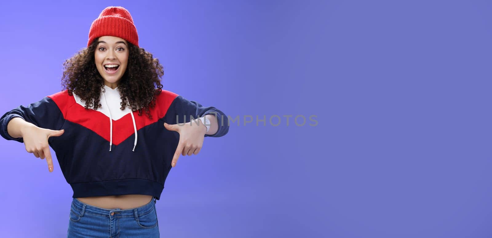 Hurry up and chek it out. Portrait of charismatic joyful and excited young cute curly-haired girl in beanie and stylish outfit pointing down, smiling broadly inviting join fun over blue background by Benzoix