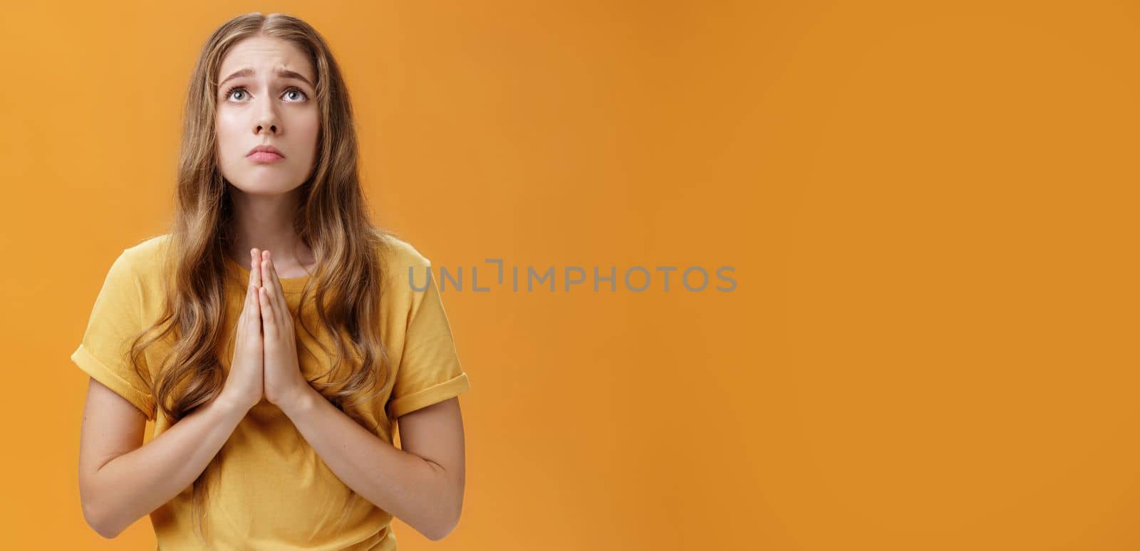 Hopeful uneasy and worried gloomy faithful girl in t-shirt holding hands in pray against chest looking up with sad look praying making wish for good well of family posing over orange wall. Body language concept