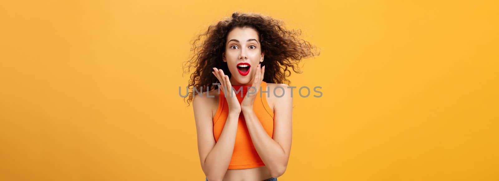 Waist-up shot of charmed enthusiastic attractive female with curly hairstyle in cropped top holding palms on cheeks and smiling astonished and surprised gazing with amazement over orange background. Copy space