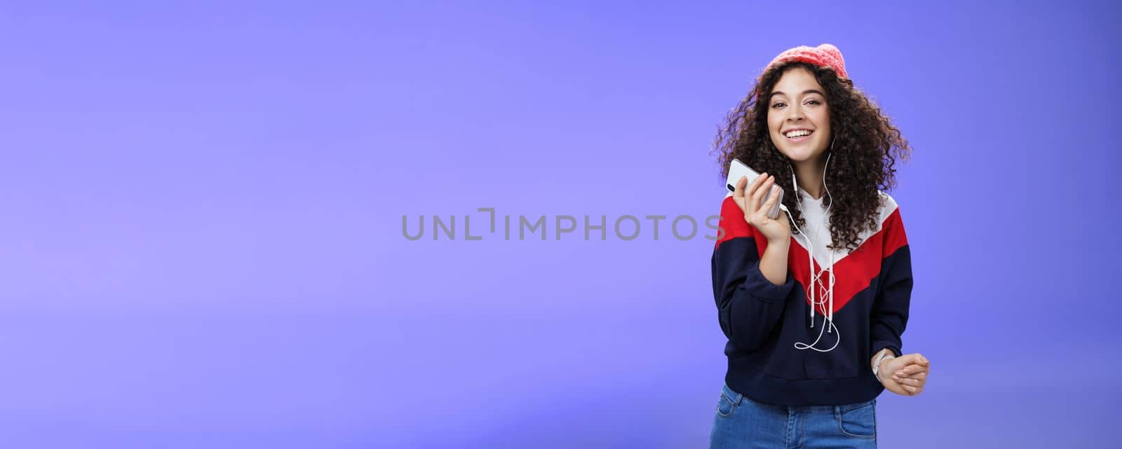 Waist-up shot of charming happy tender curly-haired female in beanie and sweatshirt listening music as dancing carefree, playing karaoke app or enjoying cool song in earphones over blue background by Benzoix