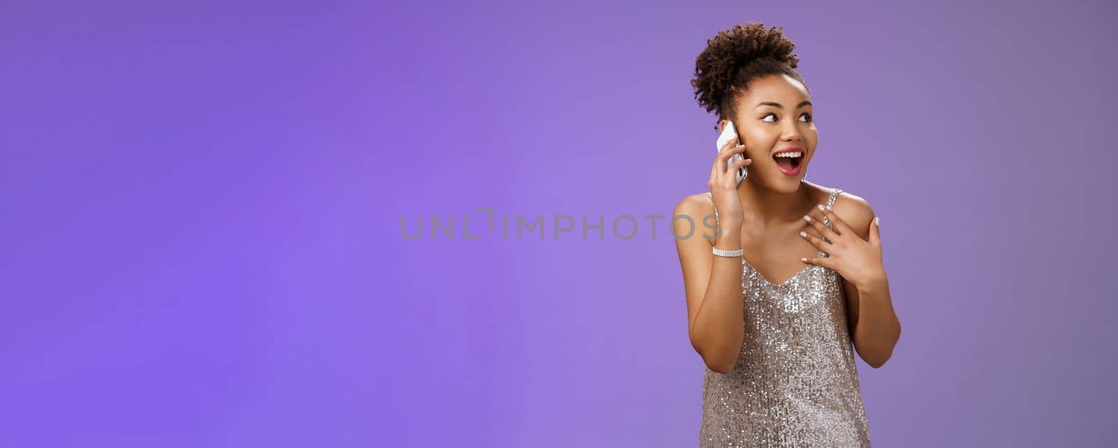 Impressed sociable african-american talkative woman in silver dress talking smartphone pointing herself amused describing emotions impressions after visit awesome party, blue background.