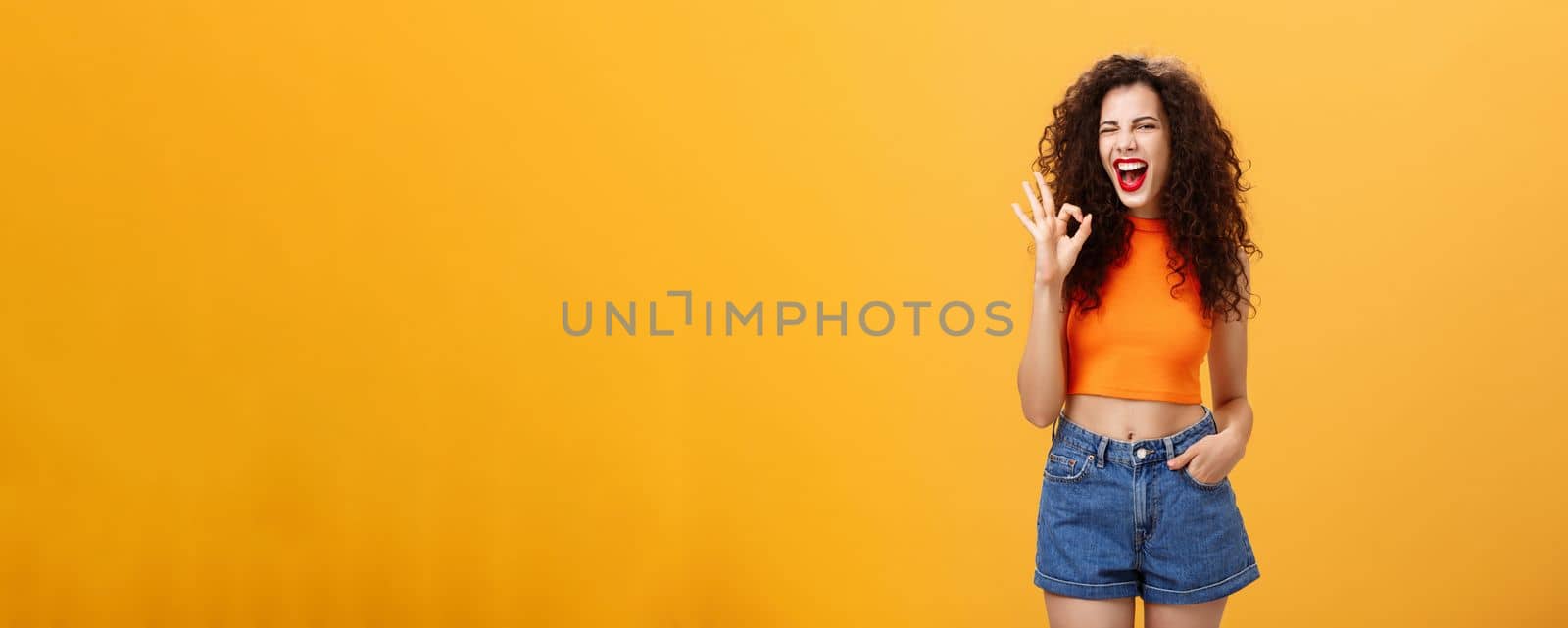 Excellent party like it. Joyful enthusiastic young curly-haired female with red lipstick in stylish cropped top winking excited smiling and showing okay or perfect sign posing over orange background by Benzoix