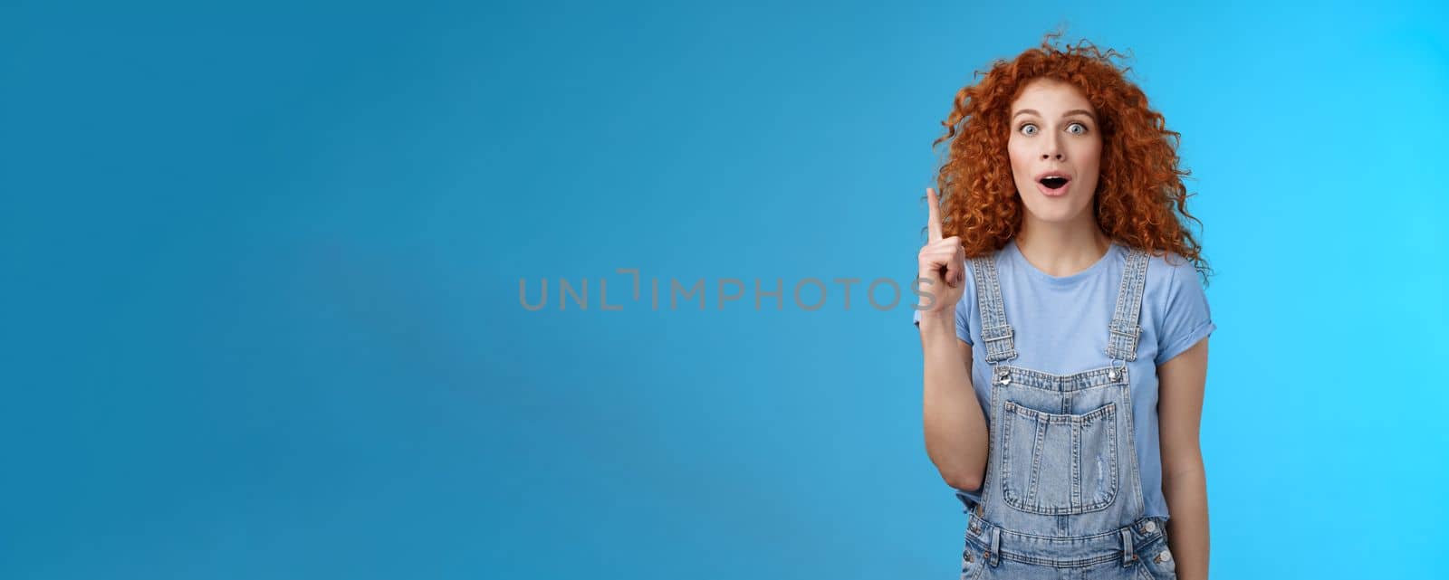 Impressed excited cute redhead curly female european student wear denim overalls t-shirt open mouth sensitive talking about amazing promo offer pointing up index finger smiling inspired by Benzoix
