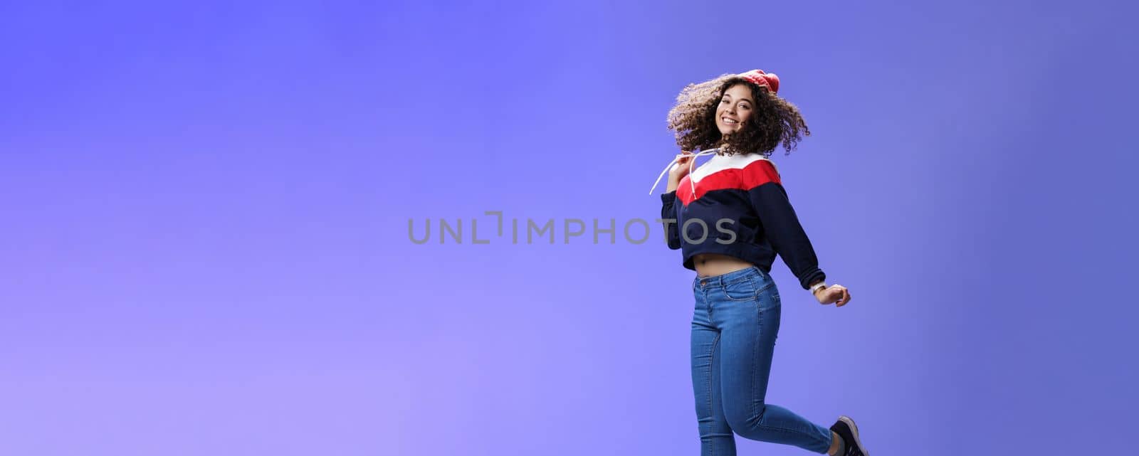 Dreamy and femenine cute girl with curly hair in winter beanie, jeans and sweatshirt jumping over blue background with satisfied carefree smile looking at camera feeling light and joyful by Benzoix