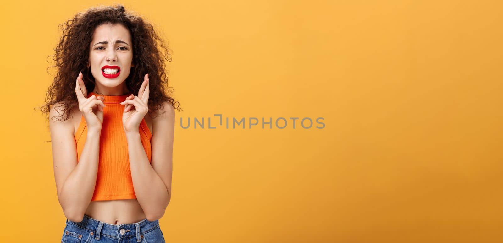 Concerned worried stylish urban girl. with curly hairstyle and red lipstick clenching teeth crossing fingers for good luck and frowning feeling nervous and anxious posing troubled over orange wall.