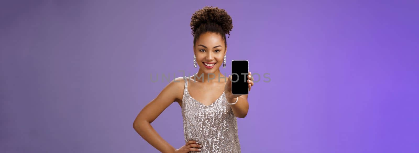 Sassy elegant african-american woman in glittering shiny silver dress hold hand waist confident pose gladly smiling extend arm showing smartphone display check out cool app blue background by Benzoix