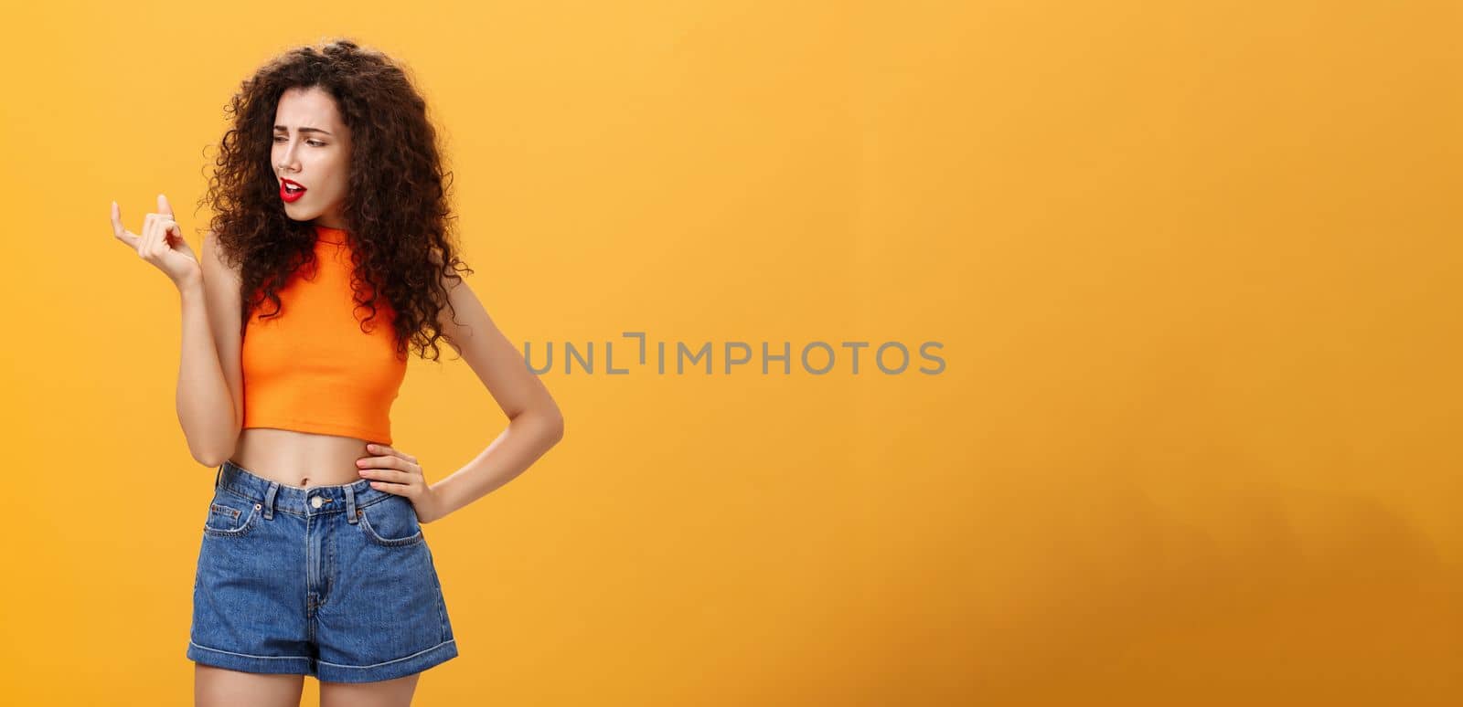 Portrait of moody arrogant and displeased european female with curly hairstyle and cropped top showing small and tiny object with fingers looking at little thing with disdain over orange background. Copy space