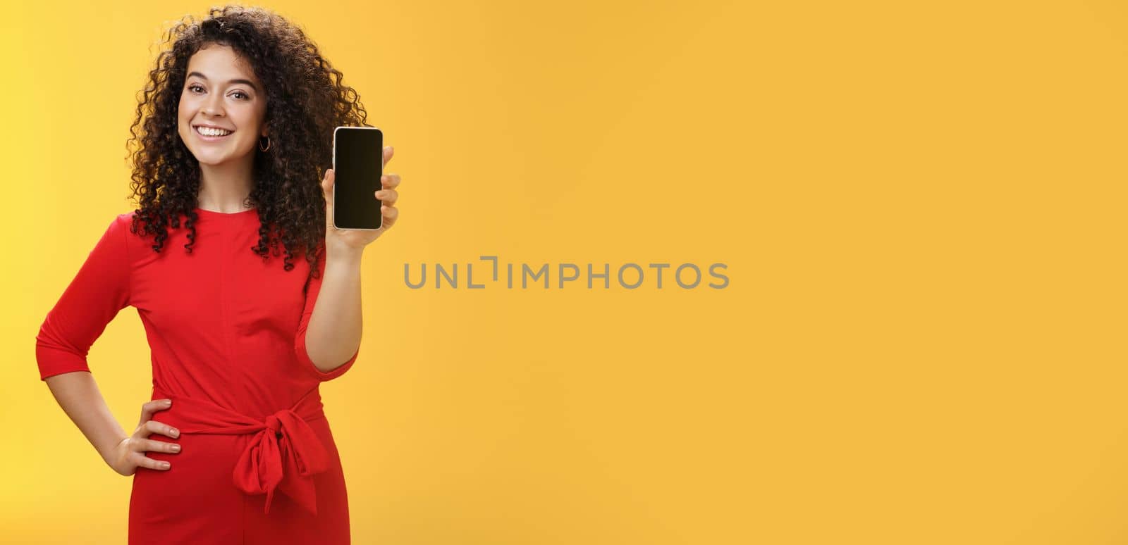 Girl brag with new phone she got on christmas feeling delighted holding mobile device in hand showing smartphone screena t camera, smiling broadly with uplifted mood over yellow background by Benzoix
