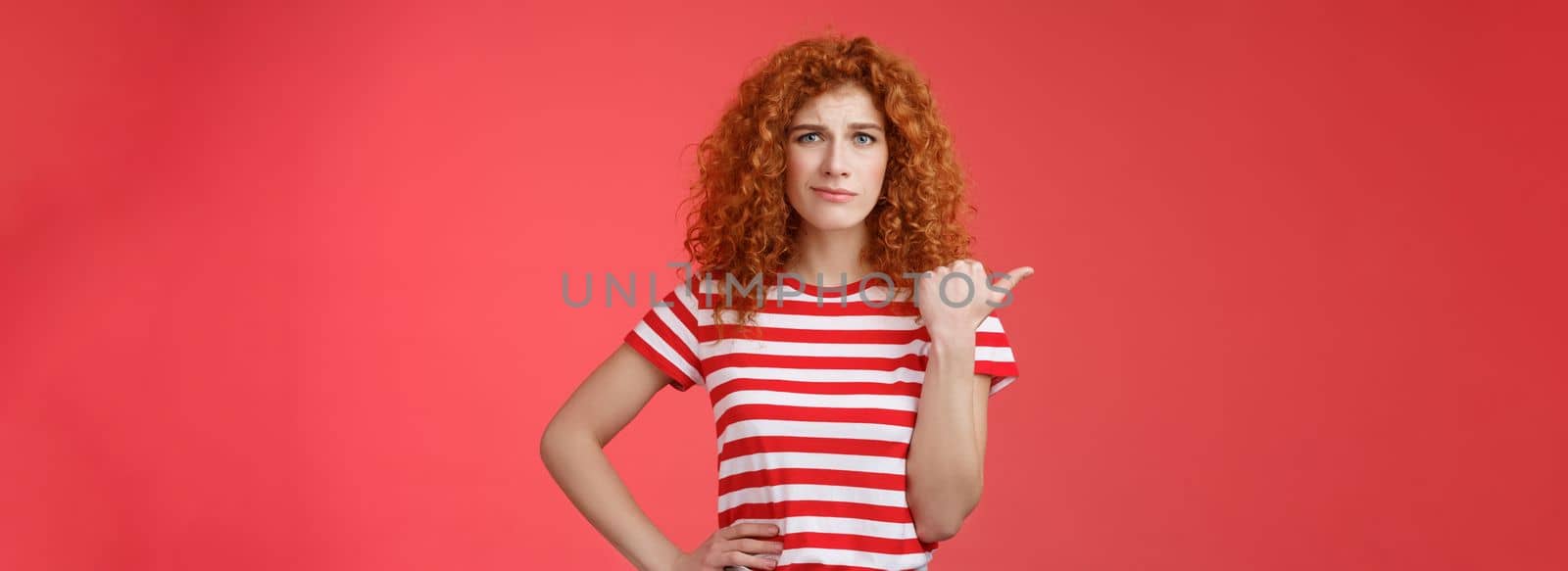 Girl not sure best choice. Doubtful redhead curly woman look suspicious frowning uncertain stare hesitant pointing left thumb unsure buy or not standing red background displeased. Copy space