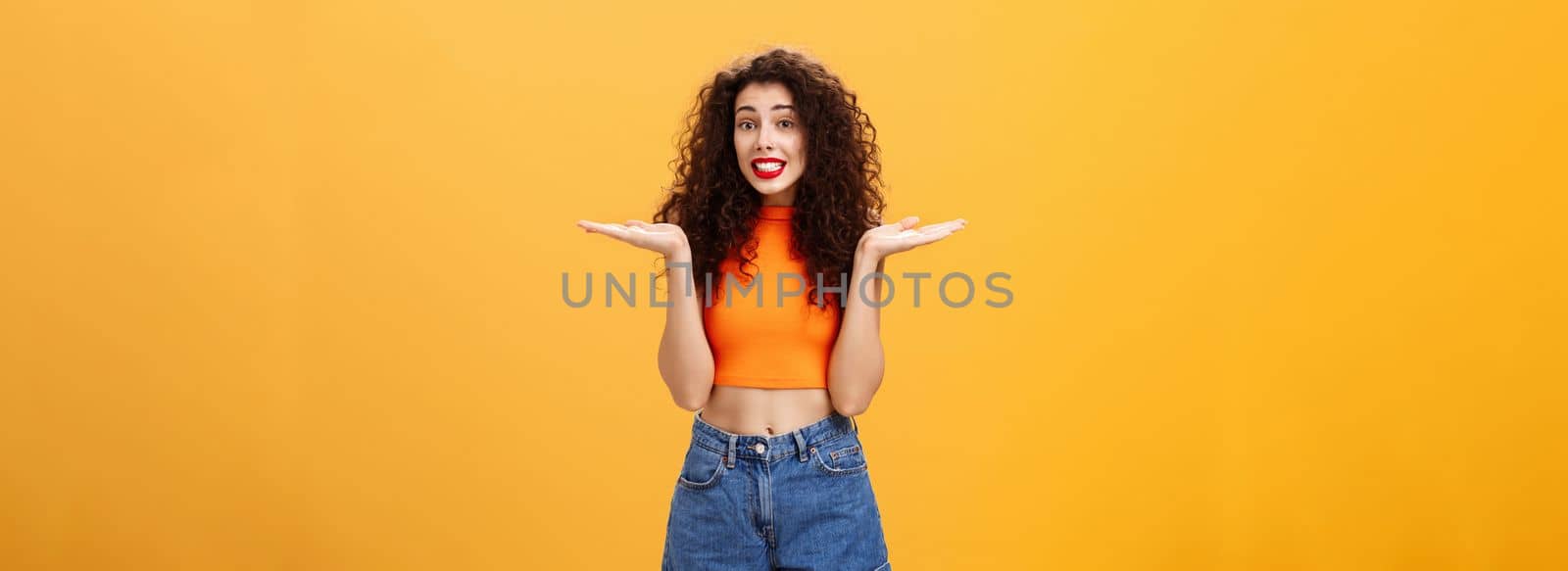 Oops honey sorry. Clueless silly stylish european female with curly hairstyle in cropped top shrugging with hands spread aside in unaware gesture smiling guilty and questioned over orange background.