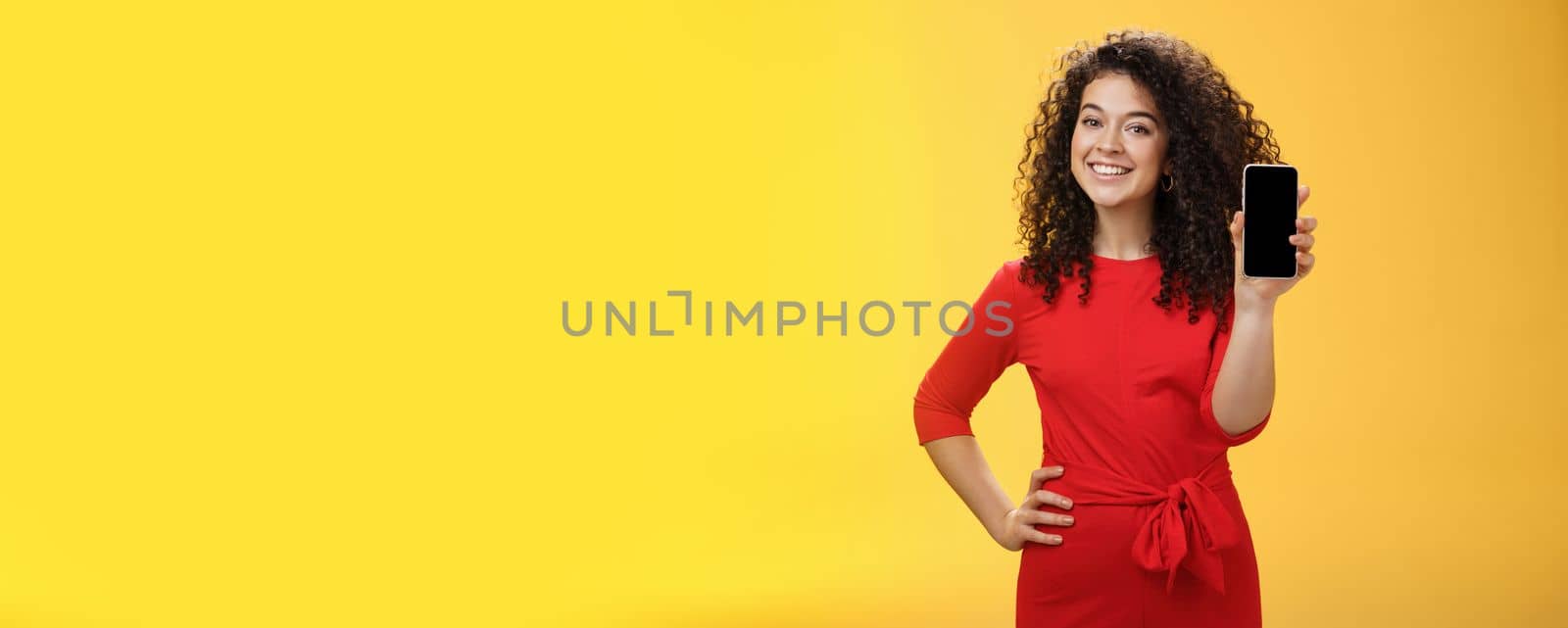 Girl brag with new phone she got on christmas feeling delighted holding mobile device in hand showing smartphone screena t camera, smiling broadly with uplifted mood over yellow background. Technology concept