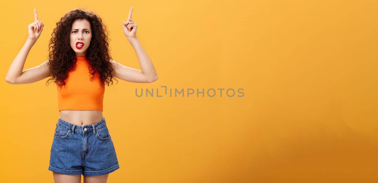 Indoor shot of displeased female client with curly stylish hairstyle red lipstick in cool cropped top pointing up demanding explanation with confused and dissatisfied expression over orange background by Benzoix