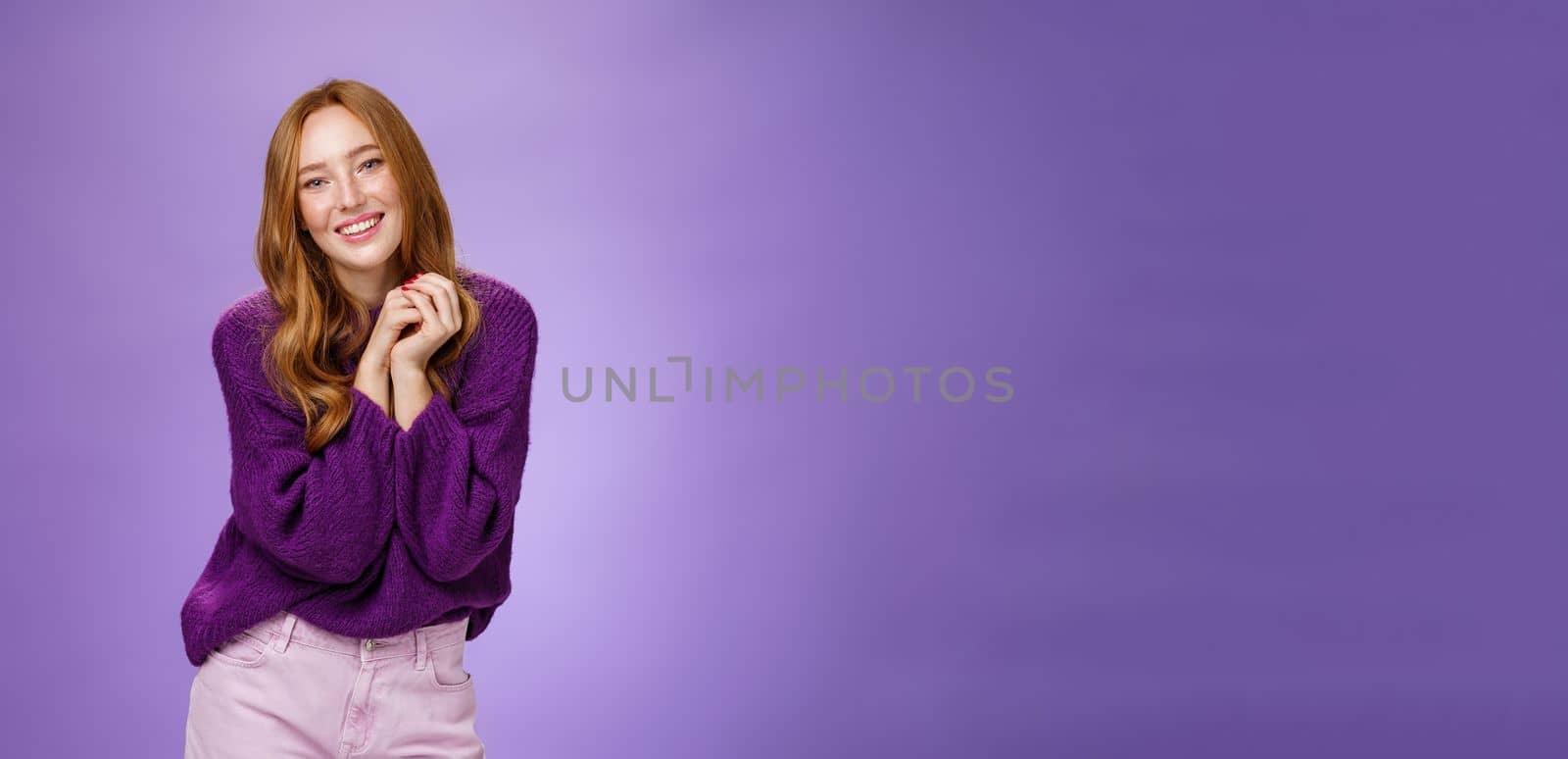 Charming and tender carefree redhead girlfriend with cute freckless holding hands together near shoulder as posing flirty and feminine smiling broadly having positive, happy attitude over purple wall by Benzoix
