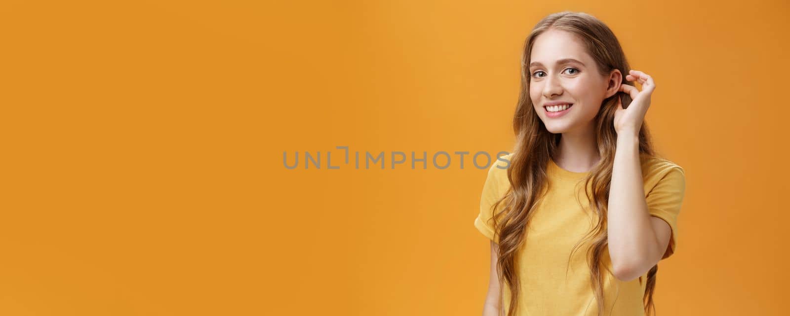 Cute and tender flirty young woman with cute wavy hairstyle flicking hair strand behind ear gazing at camera with timid sensual smile glancing with admiration at guy over orange background by Benzoix