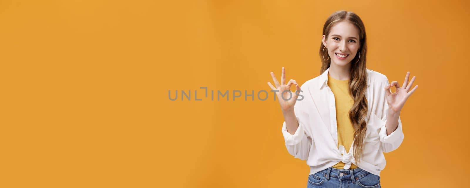 Indoor shot of assertive and assured charming stylish woman in blouse over t-shirt and accessories showing okay gesture with delighted self-assured smile giving being ok against orange wall by Benzoix