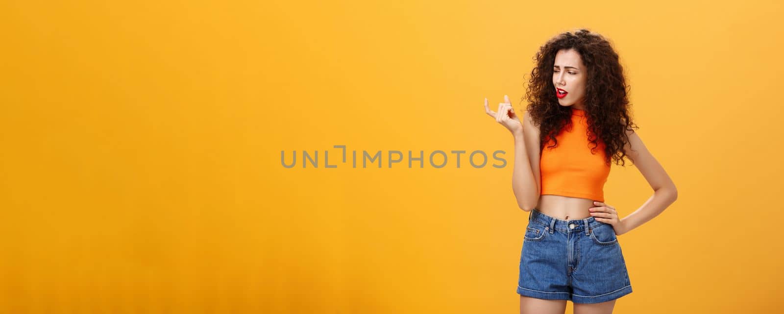 Portrait of moody arrogant and displeased european female with curly hairstyle and cropped top showing small and tiny object with fingers looking at little thing with disdain over orange background by Benzoix