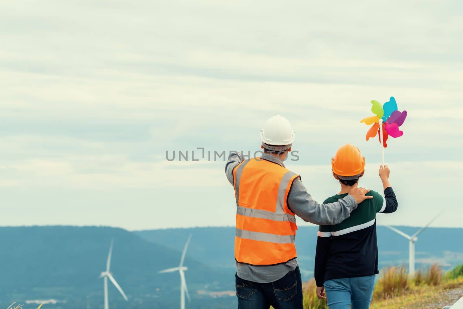 Engineer with his son holding windmill toy on a wind farm atop a hill or mountain. Progressive ideal for the future production of renewable, sustainable energy. Energy generated from wind turbine.