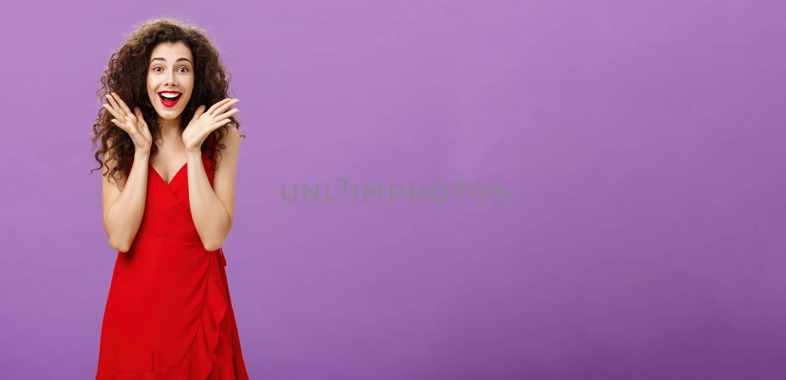 Amazed and surprised attractive charming adult. woman with curly hairstyle in red elegant dress raising palms in amazement smiling and gazing at camera astonished with charming gesture of boyfriend.