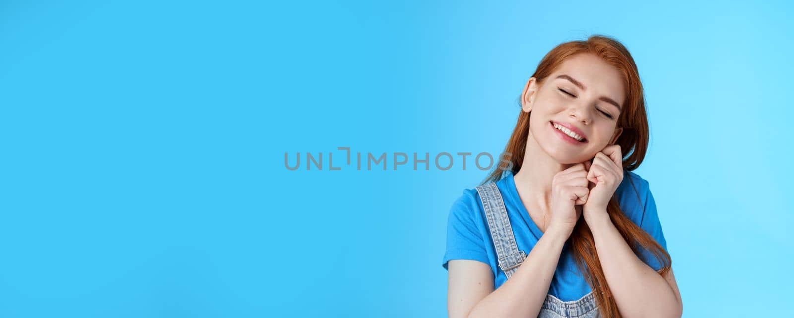 Glad cute tender redhead female, tilt head dreamy lean palms, close eyes and smiling lovely, daydreaming, recall nice memories, cherish moments, stand silly blue background nostalgic mood by Benzoix