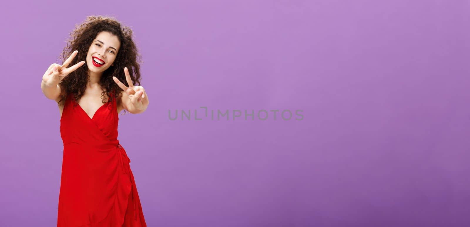 Friendly-looking peaceful european female with curly haircut in elegant red dress showing peace or victory signs, smiling broadly saying her enjoying awesome party in friends circle over purple wall by Benzoix
